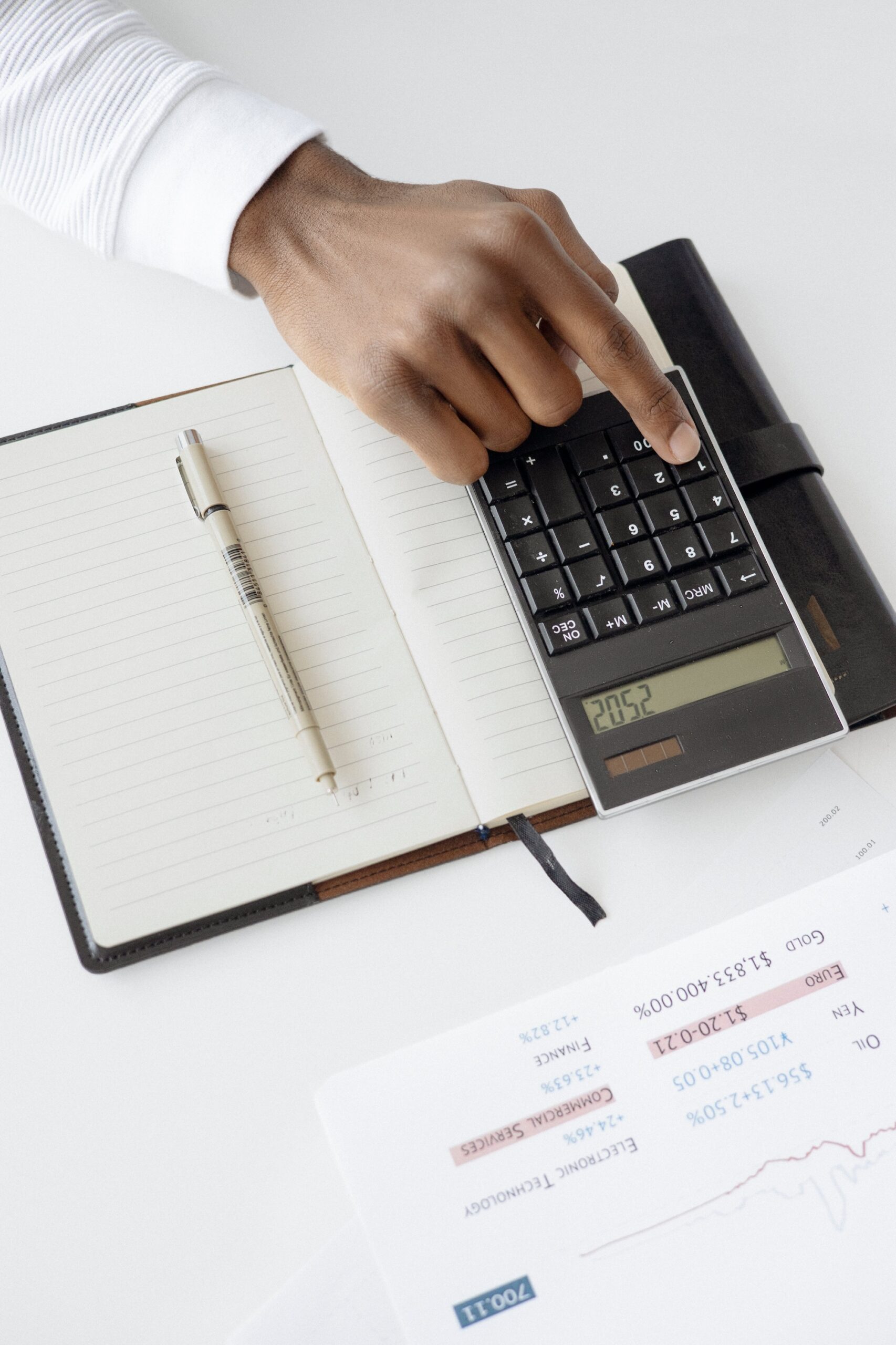 Stock photo of a hand pressing buttons on a calculator. The calculator is on the inside of an open notebook. A pen is on the other side of the inside of the notebook.